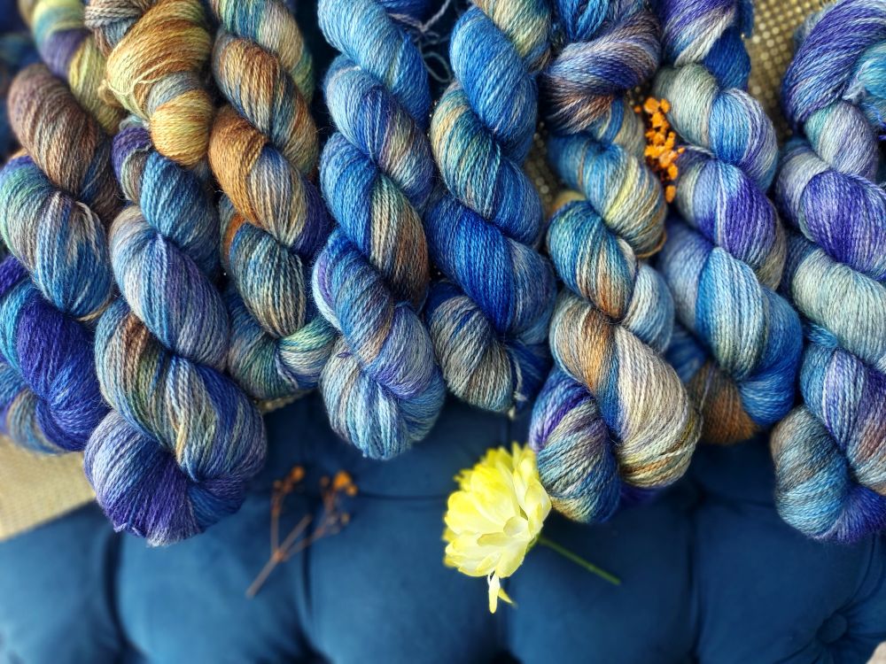 Blue, orange, indigo, soft yellow skeins with faint green in a row. There are orange and yellow dried flowers scattered 