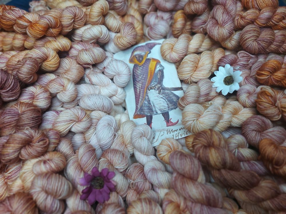 Variegated plum and bronze miniskeins, lined up in a circle around a photo of a manuscript depicting a Black nobleman, and adorned with purple and white flowers.