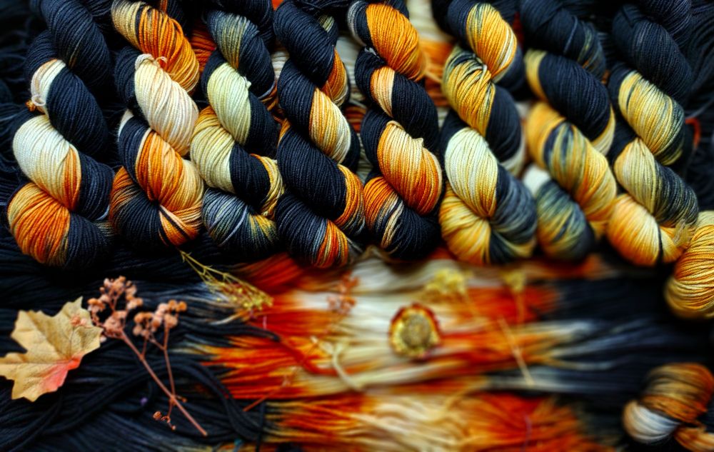 A line up of yarn skeins made in a palindrome style that is a saturated black that transitions to a deep persimmon orange and golden tip. 

There are fall leaves and dried flowers scattered around 