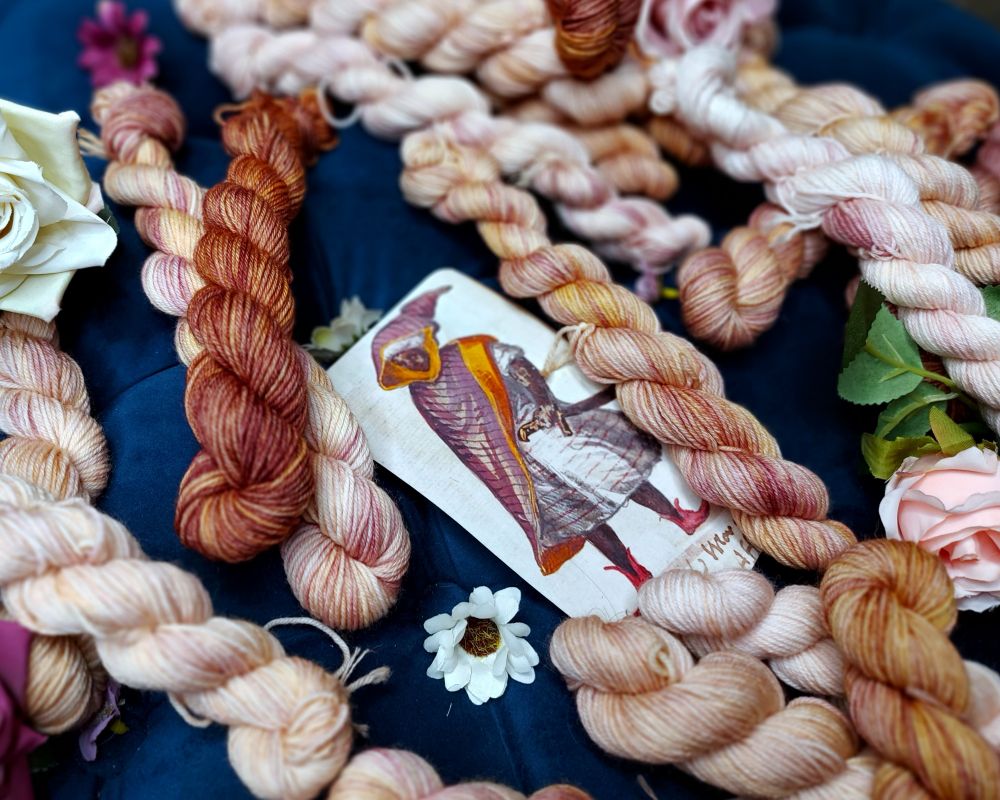 Variegated plum and bronze miniskeins, laid around a photo of a manuscript depicting a Black nobleman, and adorned with purple and white flowers.