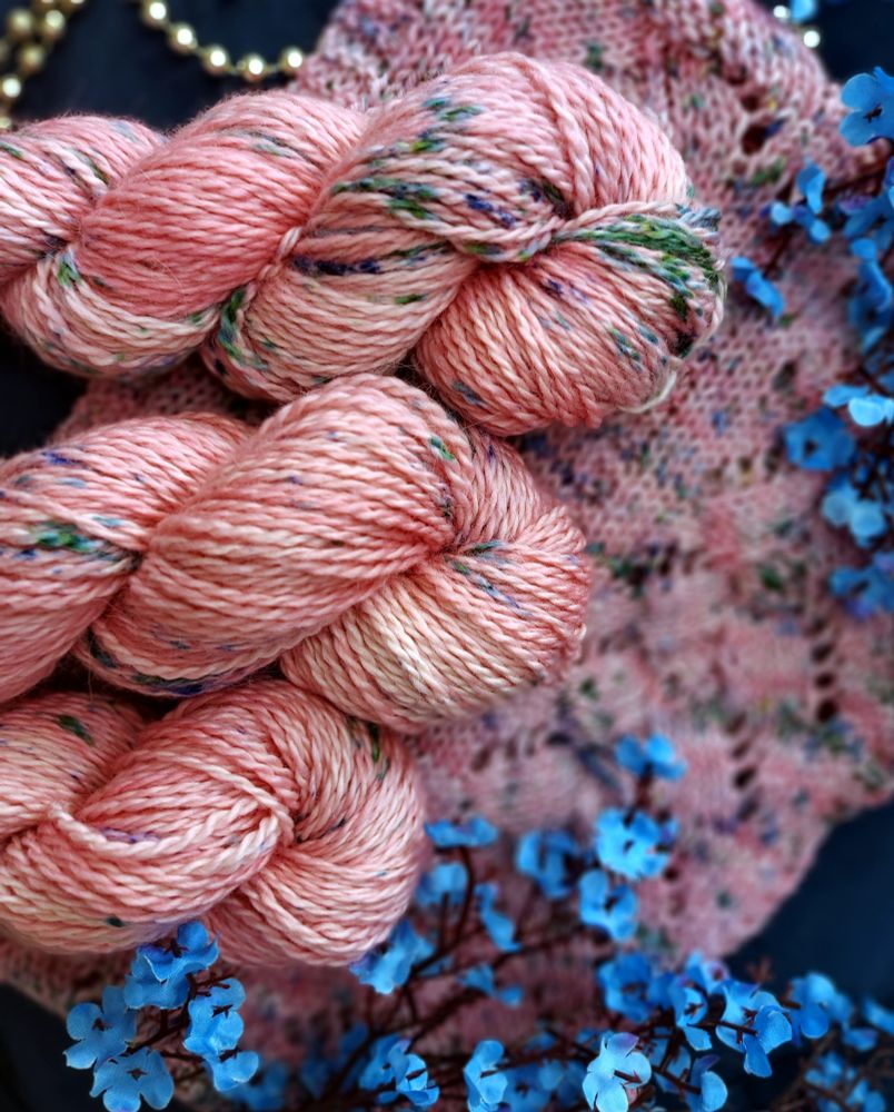 Aran weight yarn skeins that are variegated soft pink with green, and blue specks. In the background is a knit piece in the same colorway. Blue flowers are scattered around 