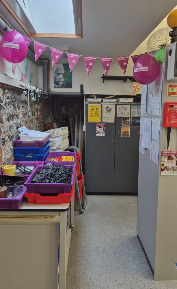 The entrance way into a prep room. To the right is the teacher's area and whiteboard. To the left is a small bench piled with trays of science equipment ready to be distributed. Across the top of the entrance way is purple bunting and balloons.