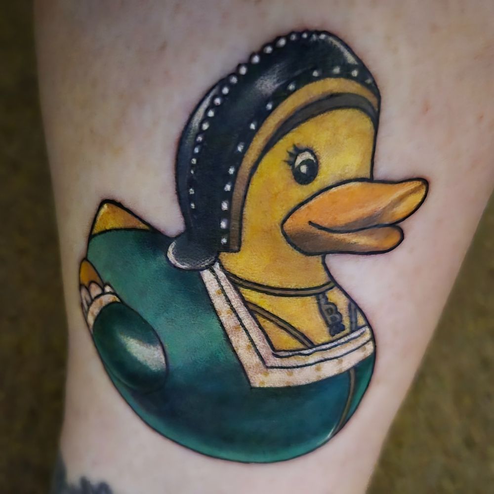A tattoo on my ankle, featuring a rubber duck dressed as Anne Boleyn. She is wearing Anne's iconic B necklace, her dress is green and she has a French hood covering her hair.