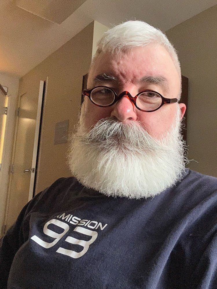 An old gay with white hair and Santa beard wearing small reading glasses.