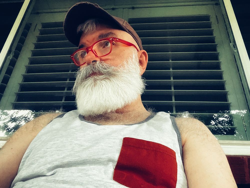 Old gay man with a Santa beard, red reading glasses, wearing a tank top sitting on a porch in front of living room windows.