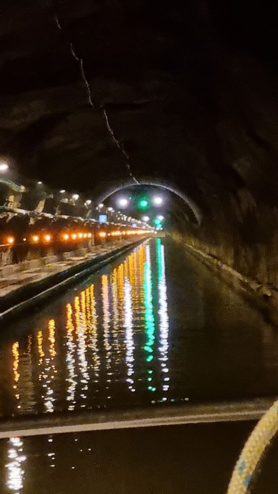 Langer Tunnel eines Wasserkanals mit Hausboot mit grüner Lampe am Ende des Tunnels und jeder Menge weisser Lampen am linken Rand die sich alle imeicht die neben Kanalwasser spiegeln.