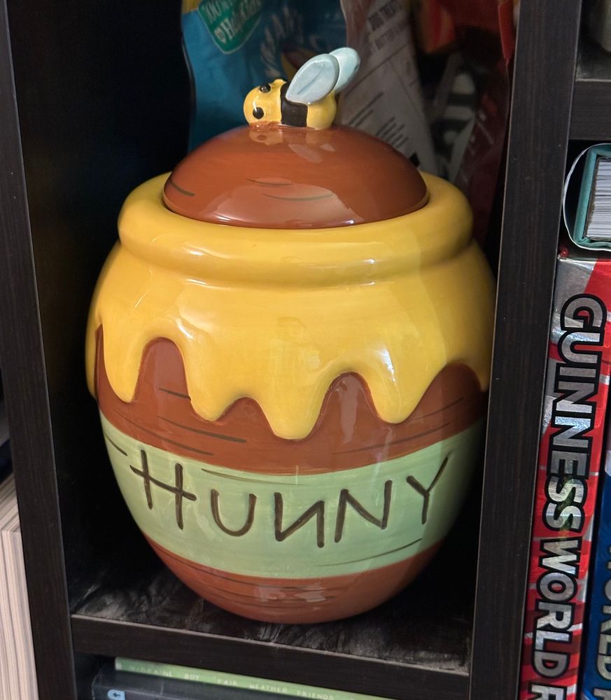 Photo of a ceramic Winnie the Pooh hunny jar with a little bee with wings sitting on top of the lid as a lid handle. It is situated in a bookcase. 