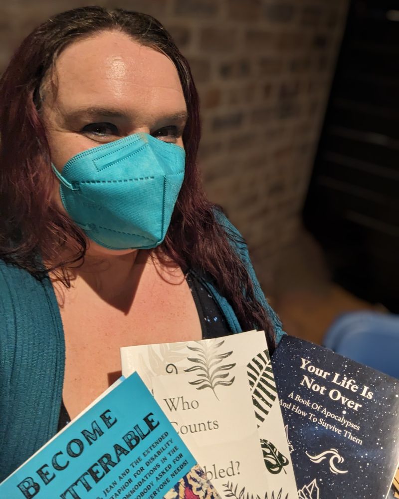 Me, a white woman with dark hair wearing a turquoise mask, holding three of my zines at a zine fair. 