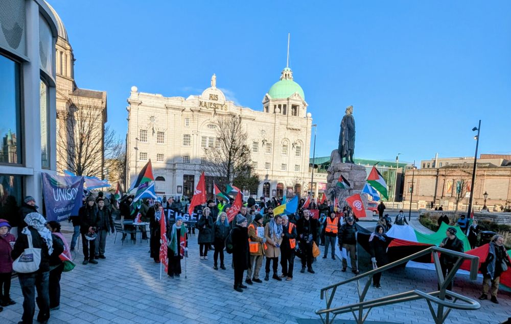 View over the courtyard above Union Terrace Gardens, showing a crowd of people with flags for trade unions, Scotland, Palestine, Sudan, and community orgs. 
