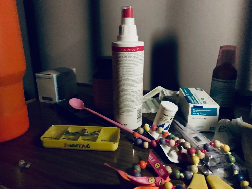 Vor einem grauen Vorhang ist ein betttisch, mit Trinkbecher, medikamentendose,Löffel, desinfektionsspray, Schmerzmittel, Fieberthermometer, Schere, und weiteren Medikamenten. 