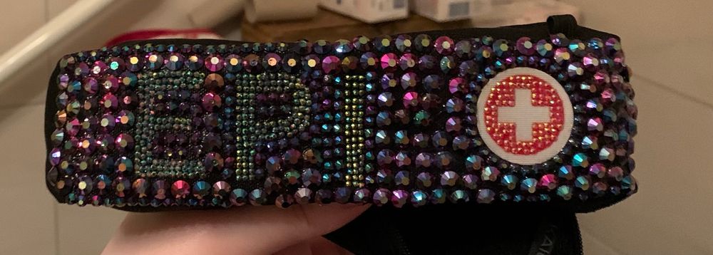 Eine schwarze etwa handgroße Tasche für einen EPI pen, beklebt mit glitzersteinen, kleine rote Steine zeigen ein Rotes Kreuz und das Wort EPI und große lila,grün, pink und blau schimmernde Steine verziehen den Hintergrund. 