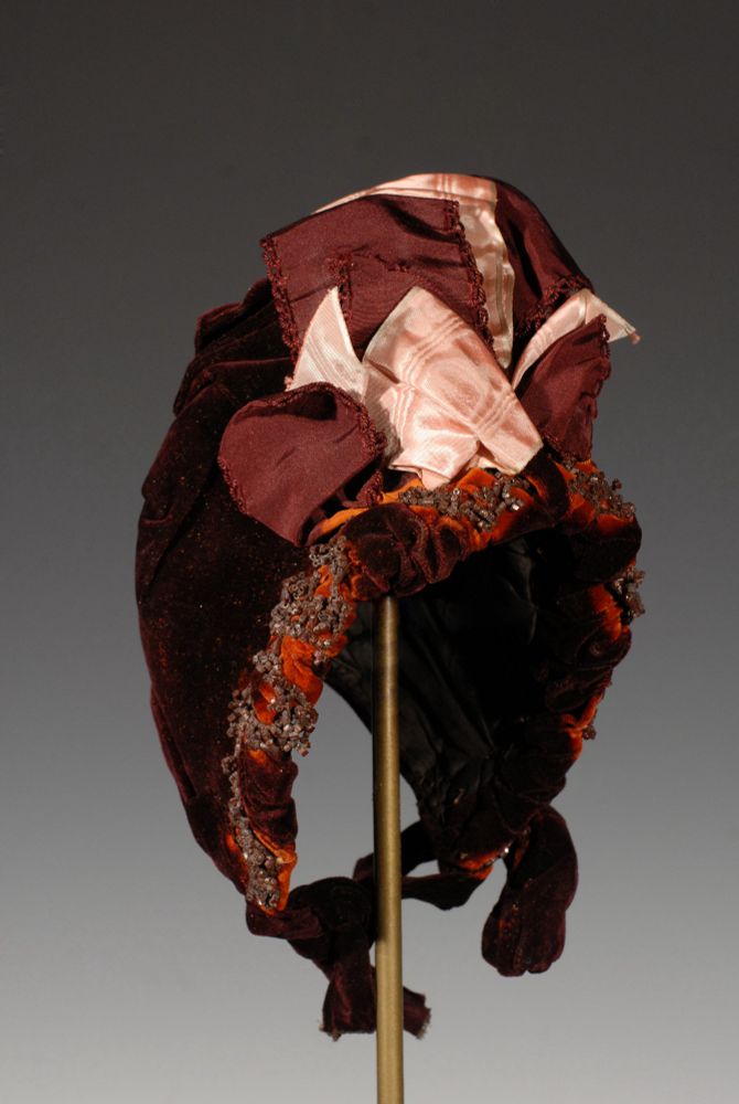 "Garnet velvet bonnet with ruched velvet brim that alternates from garnet red to rusty orange embellished with brown beads. Top of hat decorated with rose silk moire ribbon and garnet red silk faille ribbon. Bow of garnet velvet at proper left. Back of bonnet ruched."