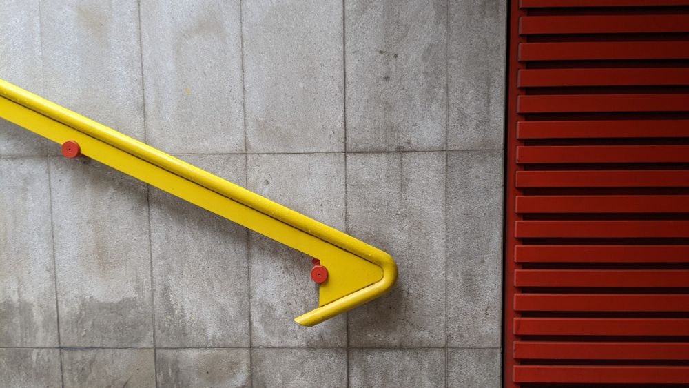on the left side of the image, a yellow banister descends diagonally along a grey stone wall, presumably follow the tread of stairs which are out of view below frame. the yellow banister is fixed to the grey wall with circular fittings painted a contrasting bright red. natural light -- perhaps from a subway entrance -- illuminates the upper left part of the image. on the right the grey stone of the wall changes to a horizontally-striped wall covering made out of metal and painted red