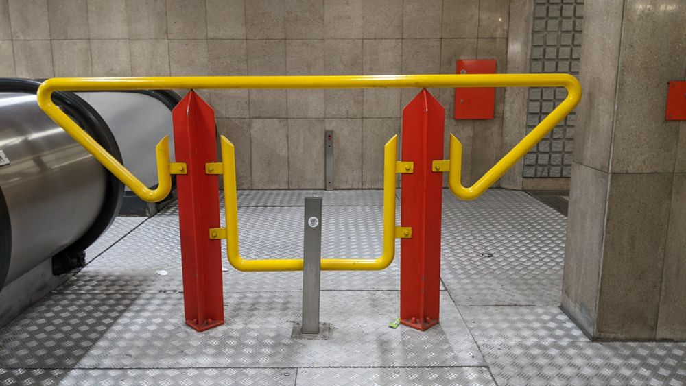 a barrier on the Milan metro. the verticals are painted red and have pointed tops; the tubular barrier itself is yellow. everything else (floor, walls) is grey -- the floor is steel tread plate, while the walls are stone (granite?)