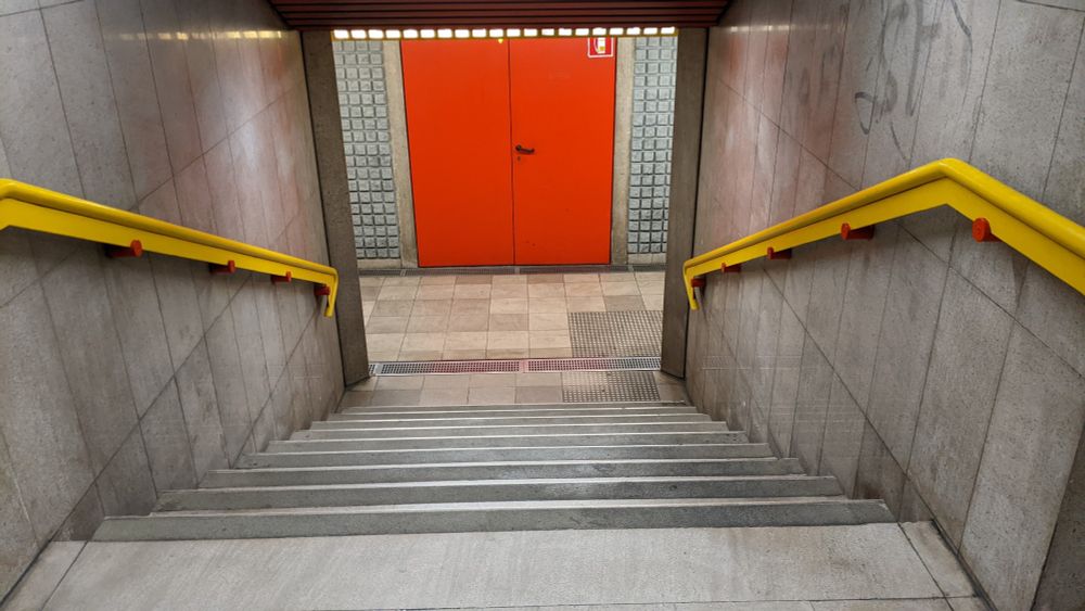a staircase. grey stone floor, grey stone walls. to left and right, on either wall, are yellow banisters fixed to the wall with circular red pins. at the bottom of the stairs is a brightly-illuminated wall, grey with a tight grid pattern, and a pair of bright red doors. above the doors is a horizontal line of small square lights