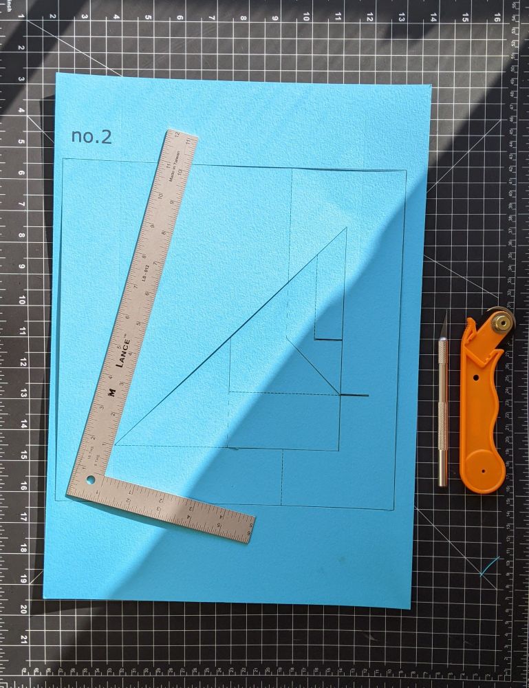 a sheet of blue paper with a metal ruler on top of a black cutting mat with a white ruled grid. next to the paper is an xacto knife and a rotary blade