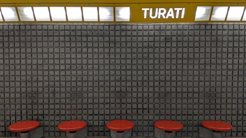 a grey wall with a grid pattern topped by a row of square lights in a long metal yellow fixture. along the base of the wall, five bright red circular seats