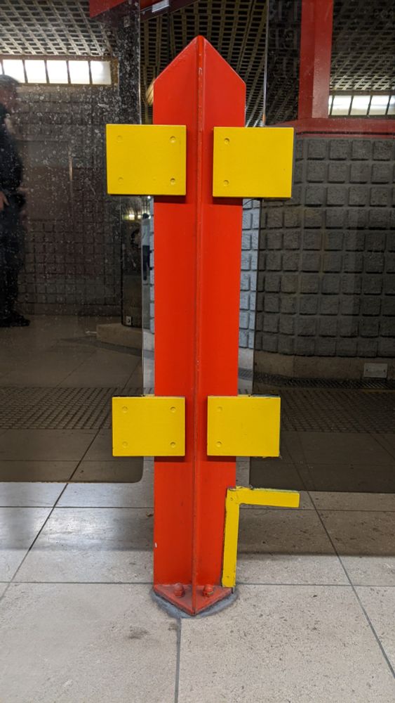 a fare barrier on the Milan metro. the barrier is made of panels of dark grey glass held up by a support post painted bright red. the support post has a cruciform plan, with the arms of the cross tapering at a forty-five-degree angle to a point at the top. the glass panels are attached to the red support post with four bright yellow rectangular panels.