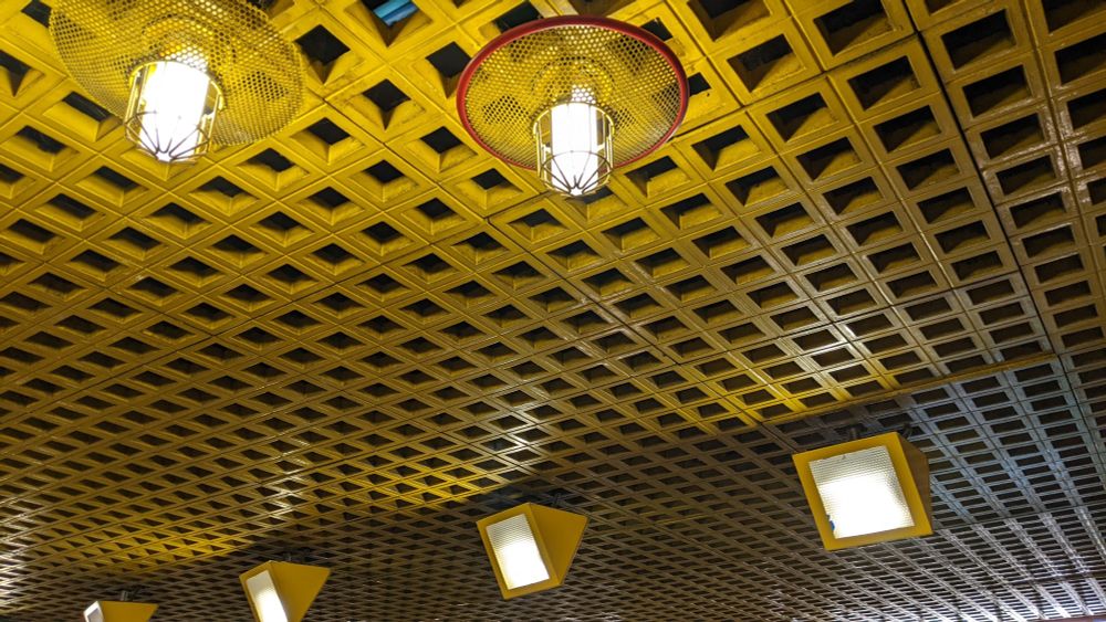 a ceiling with a square lattice painted bright yellow. the lattice is almost as deep as each square is wide -- perhaps three inches. in the foreground are two ceiling light fixtures with the bulbs set in decorative round mesh circles painted yellow (one with a red border). in the background are four triangular yellow lights, also fixed to the ceiling