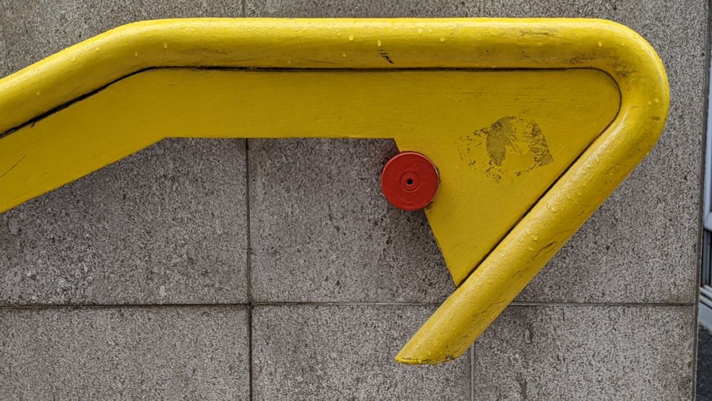 the end of a bright yellow banister, fixed to the grey stone wall behind it with a circular red pin.