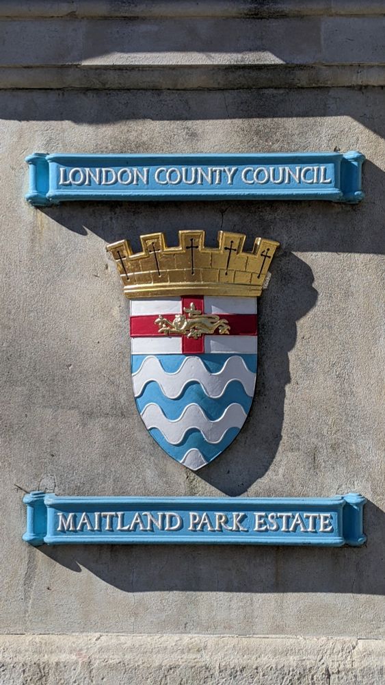 a colorful heraldic crest on a stone pillar. Above the crest, on a sky-blue scroll, the words "London County Council." Below, on a similar scroll, "Maitland Park Estate." The crest itself consists of blue and white wavy horizontal lines representing the Thames surmounted by St George's cross (red cross on white) covered by a golden lion surmounted by a golden crown