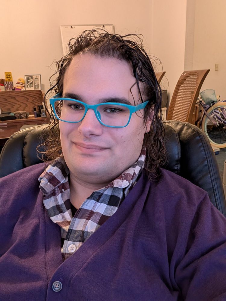 A white transfeminine person with dark curly hair sits in a black leather office chair. They're wearing a burgundy, white, and black plaid collared shirt, with a violet sweater over it, and a pair of turquoise, rectangular eyeglasses. They are smiling.