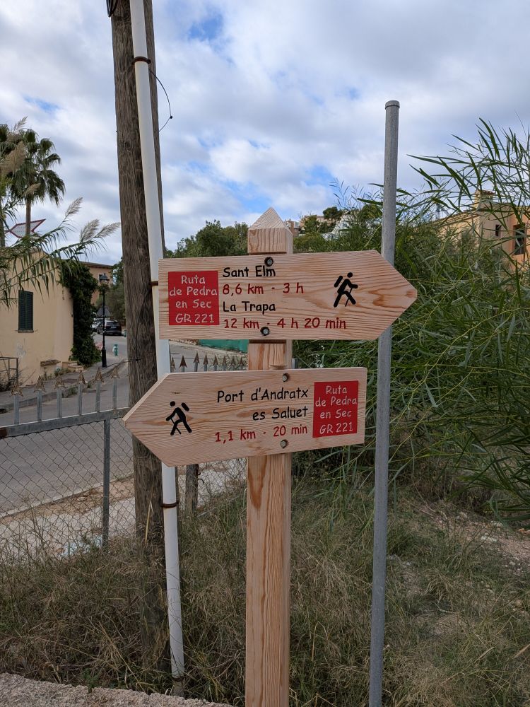 Das Bild zeigt einen hölzernen Wanderwegweiser auf Mallorca, der auf einer unbefestigten Fläche neben einem Zaun und hoher Vegetation steht.
Die Schilder weisen auf zwei Hauptziele hin:
 * Nach oben (Pfeil nach links):
   * Sant Elm und La Trapa (8,6 km, 3 Std. bzw. 12 km, 4 Std. 20 Min.).
   * Diese Route ist Teil der GR 221, der bekannten "Ruta de Pedra en Sec" (Trockenstein-Route).
 * Nach unten (Pfeil nach links):
   * Port d'Andratx es Saluet (1,1 km, 20 Min.).
   * Auch diese Strecke ist Teil der GR 221.
Im Hintergrund sind einige Gebäude, Palmen und ein hellblauer, bewölkter Himmel zu sehen.
