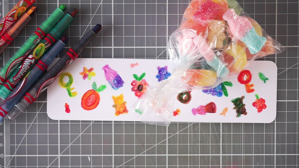 A hand-drawn paper bookmark with colourful sour candies drawn on. A bag of said sour candies is to the top right of the bookmark, and several multi-coloured swirl crayons to the top left. The background is a grid cutting mat.