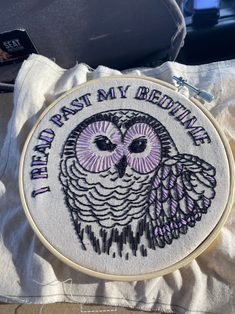 An embroidery piece in a wooden hoop on cream colored fabric. An owl in profile is outlined in black with purple details around its eyes and on its wing. Words in black and purple arc over its head saying, “I read past my bedtime.”
