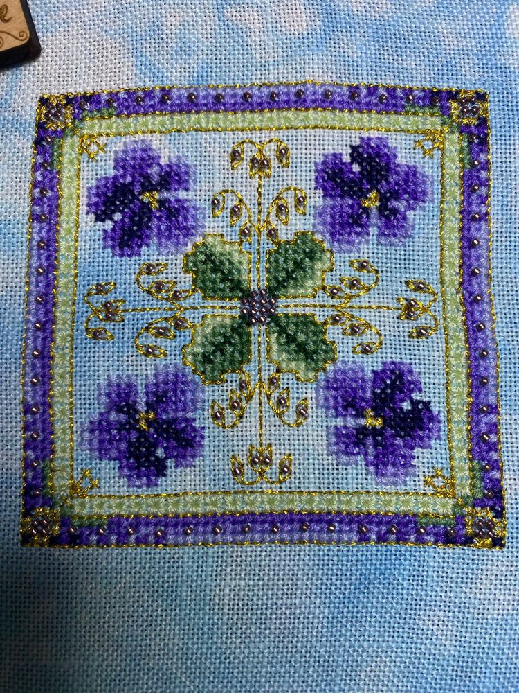 A cross stitch piece with 4 purple violets and 4 green leaves surrounded by a green, purple, and gold border. 