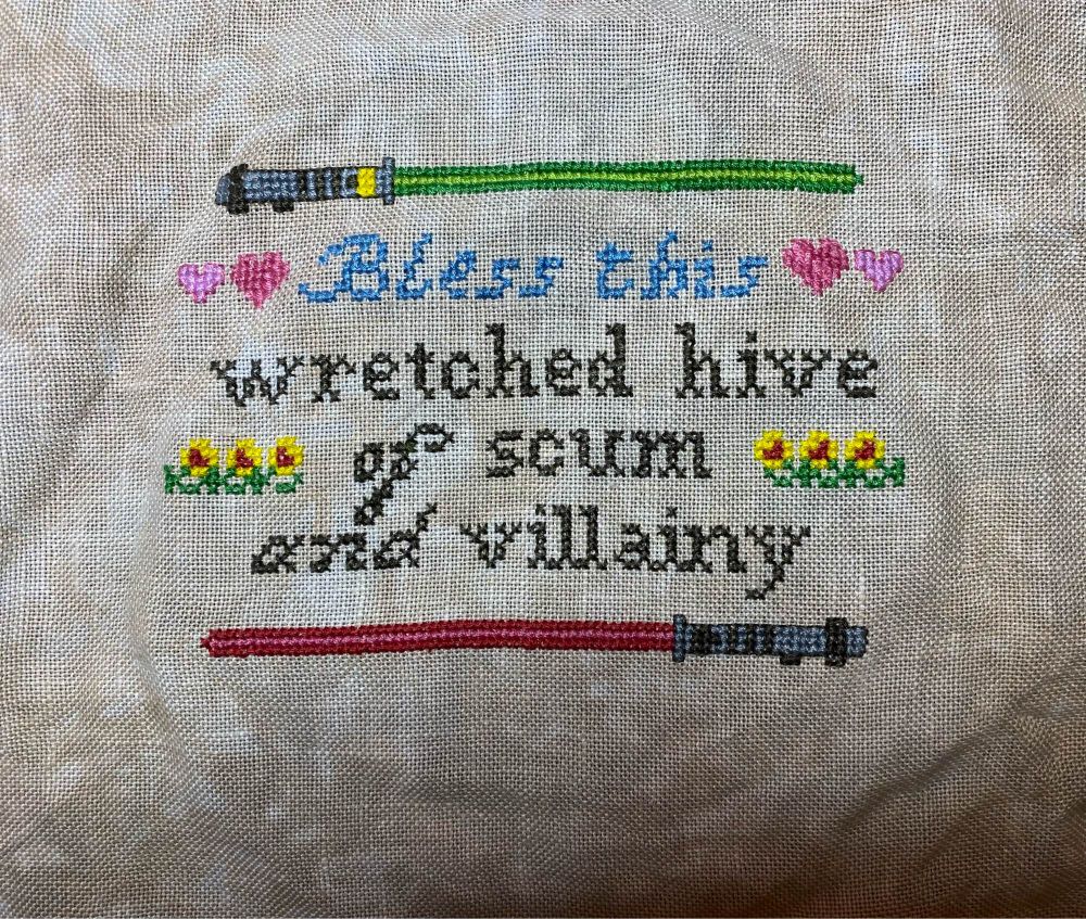 A cross stitch with two lightsabers, hearts, and flowers and the words, “Bless this wretched hive of scum and villainy.”