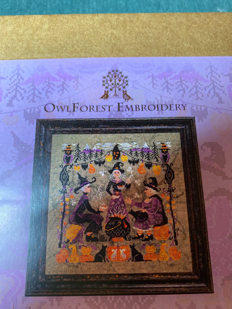 A box label for an embroidery kit shows a cross stitch piece with 3 witches seared around a cauldron surrounded by cats, owls, pumpkins, bats, and bees. 