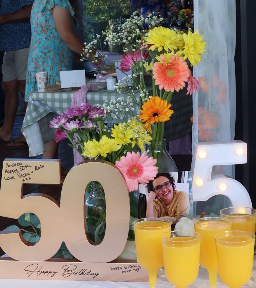 A rose gold 50 sign next to flowers in a vase and small glasses of mimosas. 