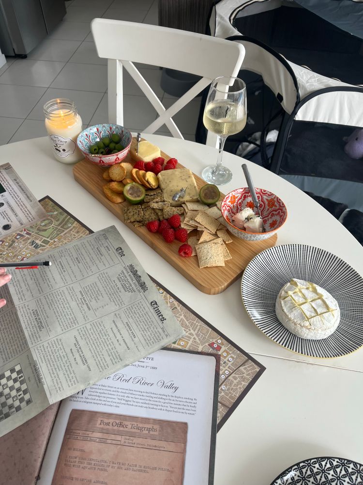 A platter with cheese , fruit and crackers on it. Some paper resembling a news paper and old book lie on the table. 