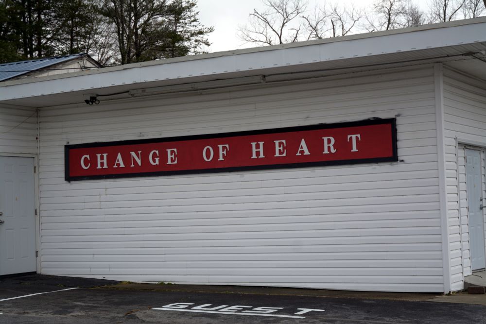 A building with white vinyl siding with a large red sign, with black outline reading "CHANGE OF HEART" in white lettering.