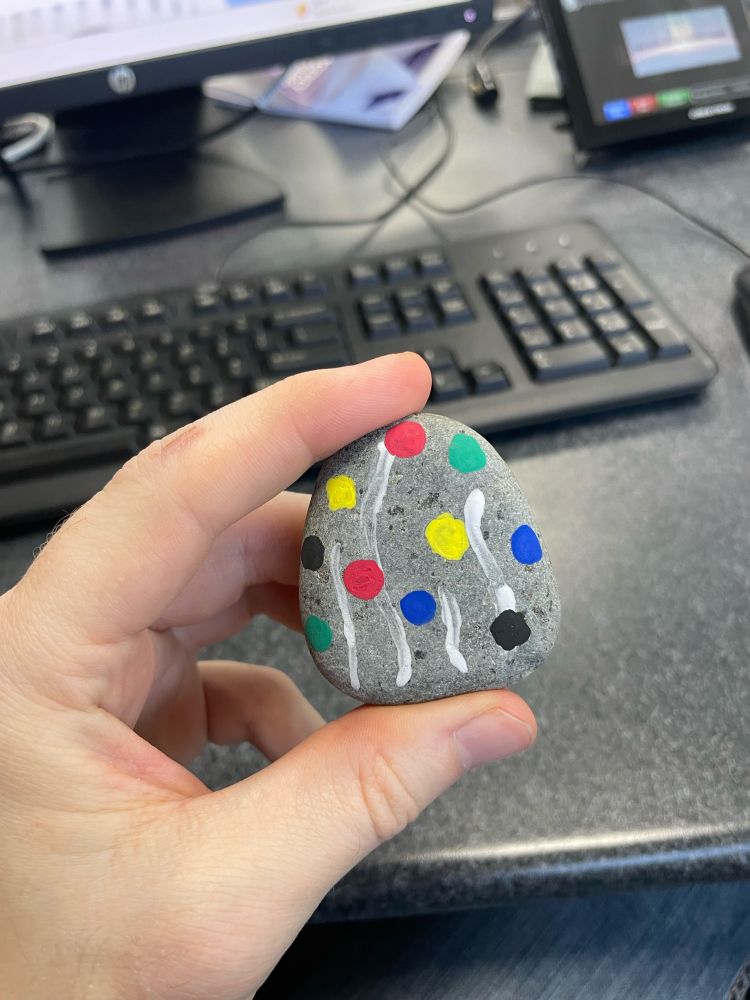 A hand holding a flat, smooth stone with polka dots and slightly wavy lines painted on it. 