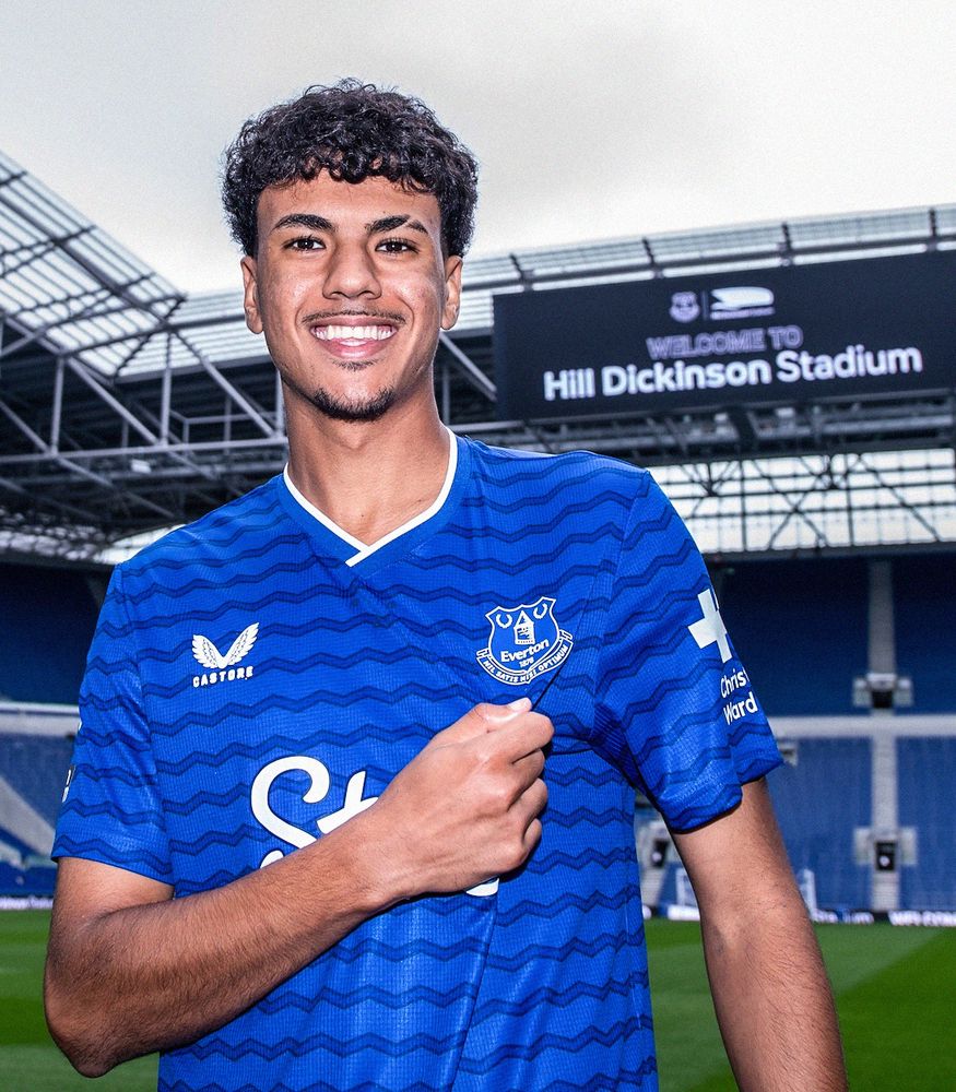Adam Aznous wearing a blue Everton shirt, stood in the middle of their brand-new stadium. He's looking at the camera and smiling.
