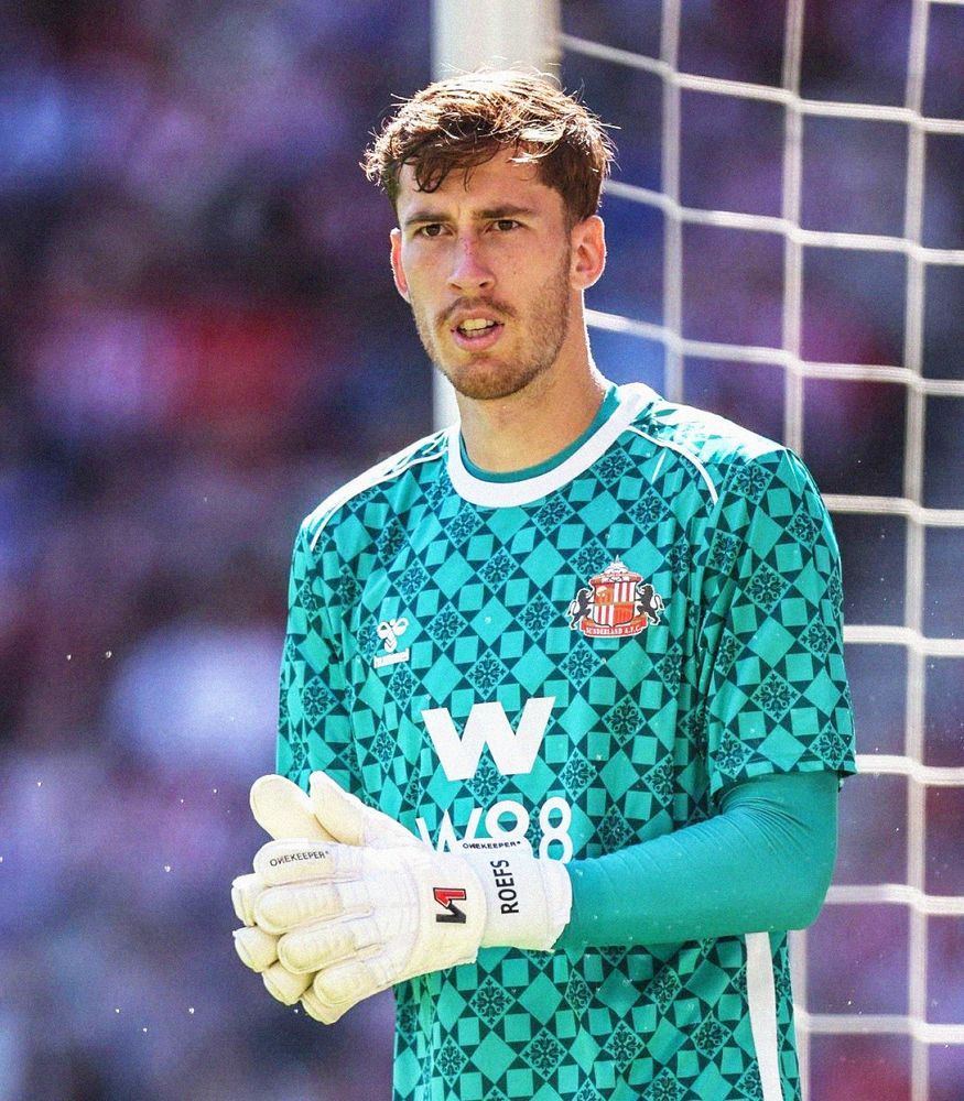 Close-up photo of Robin Roefs in a turquoise Sunderland goalkeeper shirt, clapping his hands together. In the background is a blurred crowd and a partial goal net.