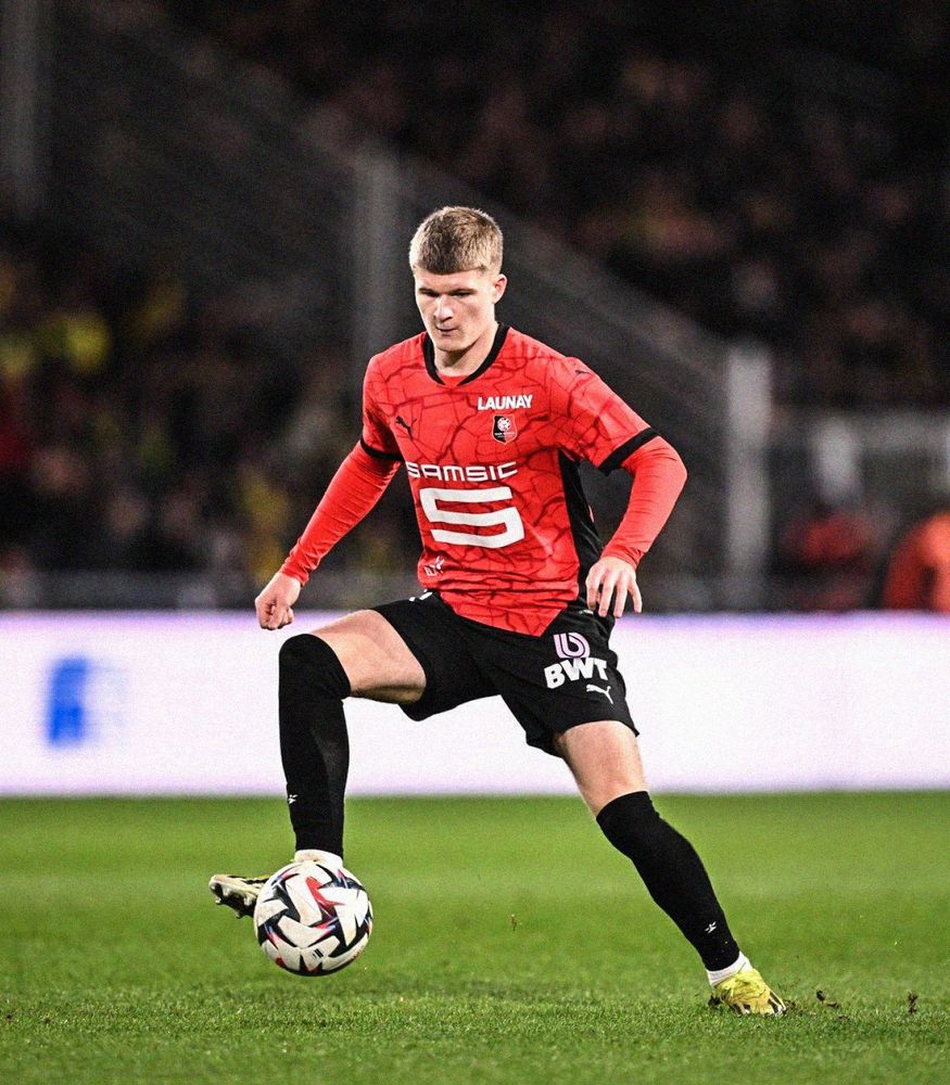 Jordan James controlling a football with his right foot wearing a red Stade Rennais shirt with black shorts and black socks. 
