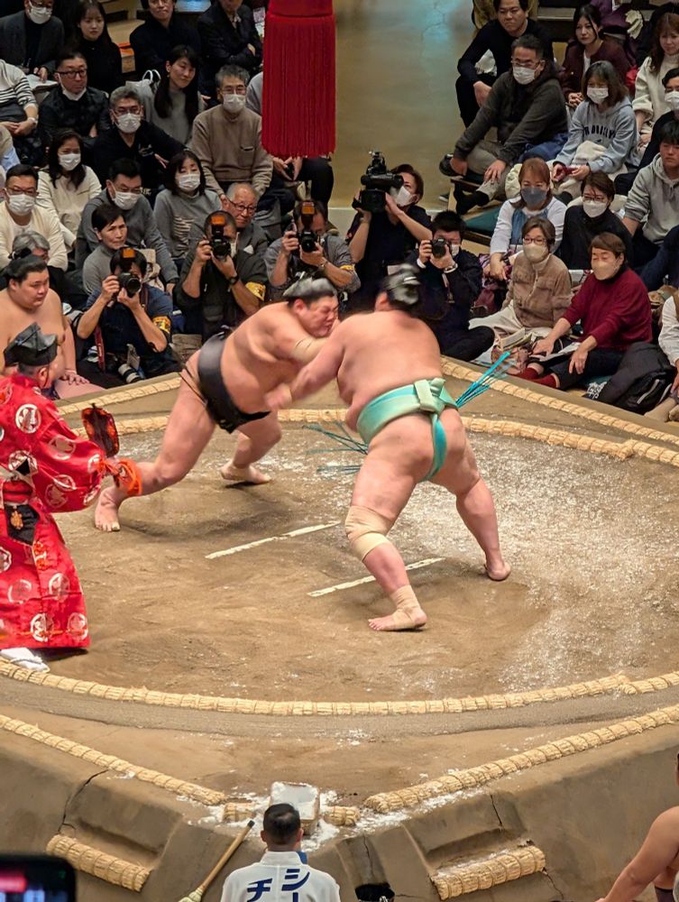 Two Rikishi launch into battle. One is wearing a black mawashi and the other a light blue.