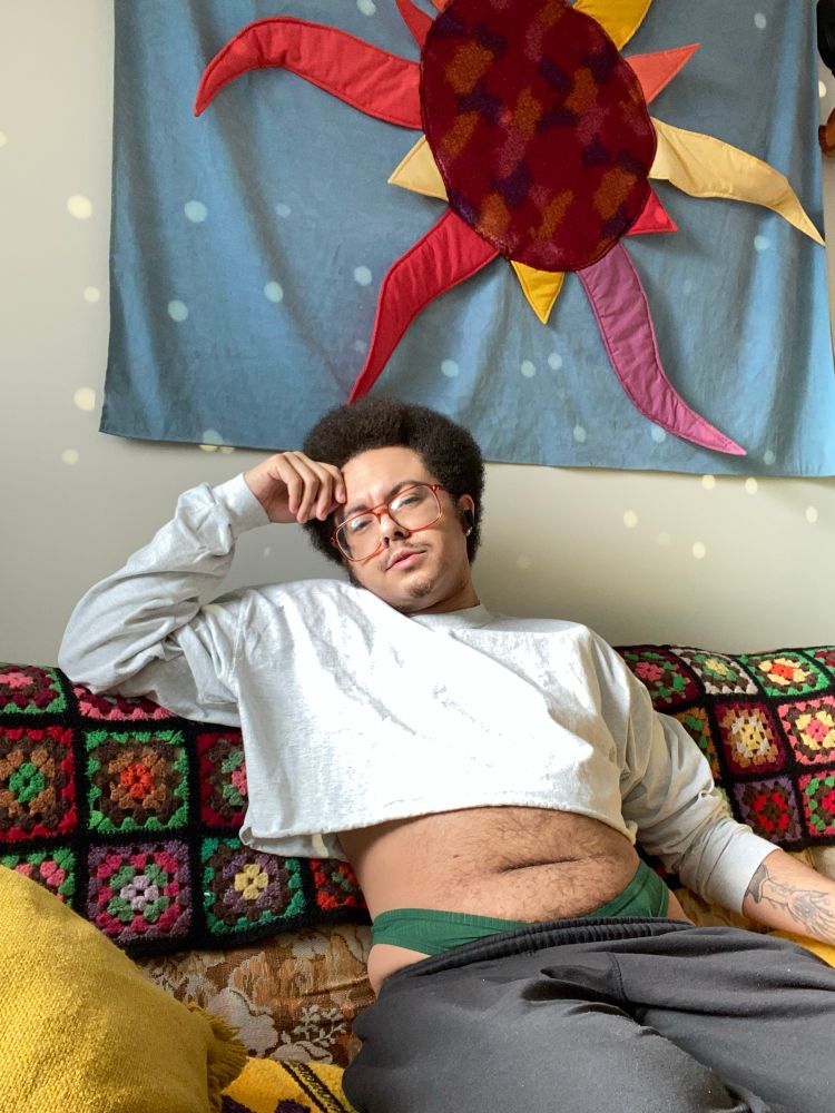 jamie is reclined on the couch, looking at you dreamily. he’s wearing a light grey long sleeve crop top, and has a kelly green thong peeking out of a pair of black sweatpants. behind him is a tapestry of a sun and the wall is covered in spots of light from an unseen mirror ball.