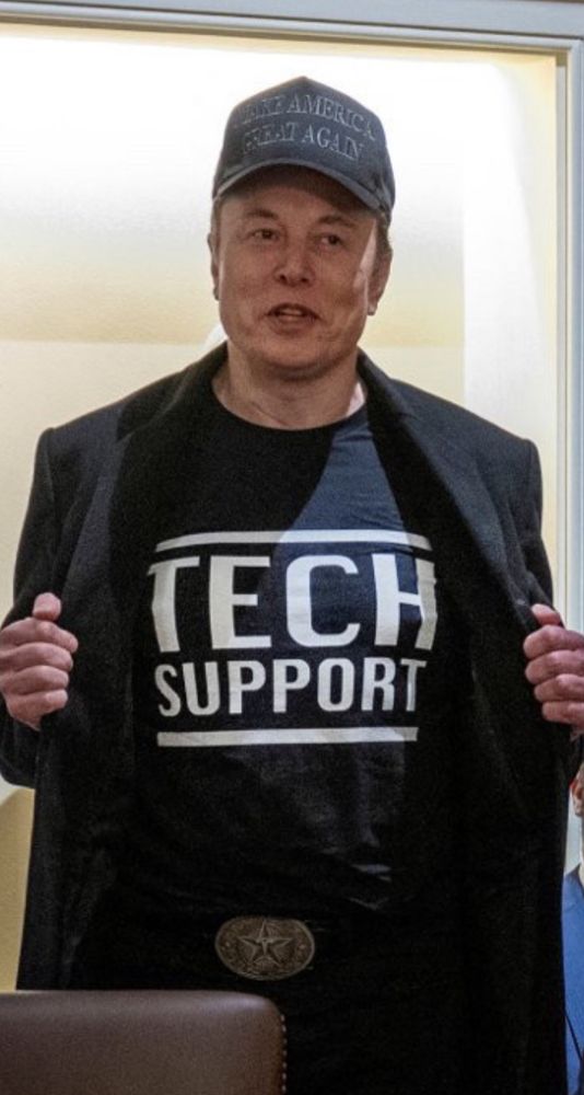 Elon Musk mit schwarzem T-Shirt mit der Aufschrift „TECH SUPPORT“ in weißen Buchstaben. Darüber trägt er eine schwarze Jacke, die er leicht geöffnet hält, um das T-Shirt zu zeigen. Er trägt außerdem eine lächerliche dunkle Baseballkappe mit der Aufschrift „MAKE AMERICA GREAT AGAIN“. Sein Gürtel hat eine auffällig hässliche Schnalle mit einem Sternmotiv. Der Hintergrund zeigt einen Innenraum mit warmem Licht.