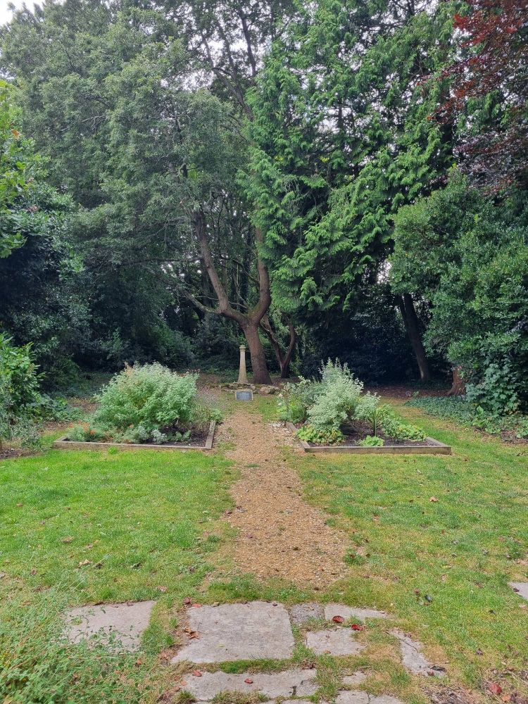 Path between flowerbeds leading to small lighthouse-shaped sculpure, surrounded by trees and grass