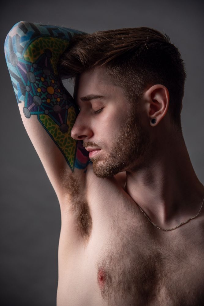 He looks to the left, nose resting near his armpit with a colorful tattoo’s arm reaching behind his head. 