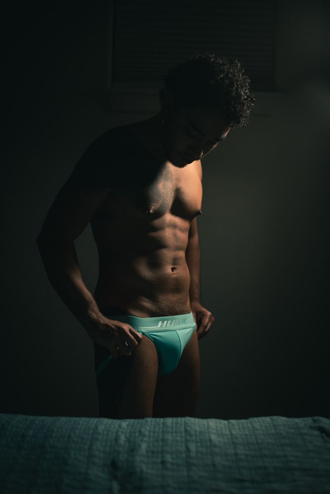 He stands, bedside, in a dimly lit room. Just beginning to tug at the leg straps of his turquoise jock strap, he looks to the camera - softly lit from above and right. His abs and chest are on display, a tattoo covering one of his pecs. 
