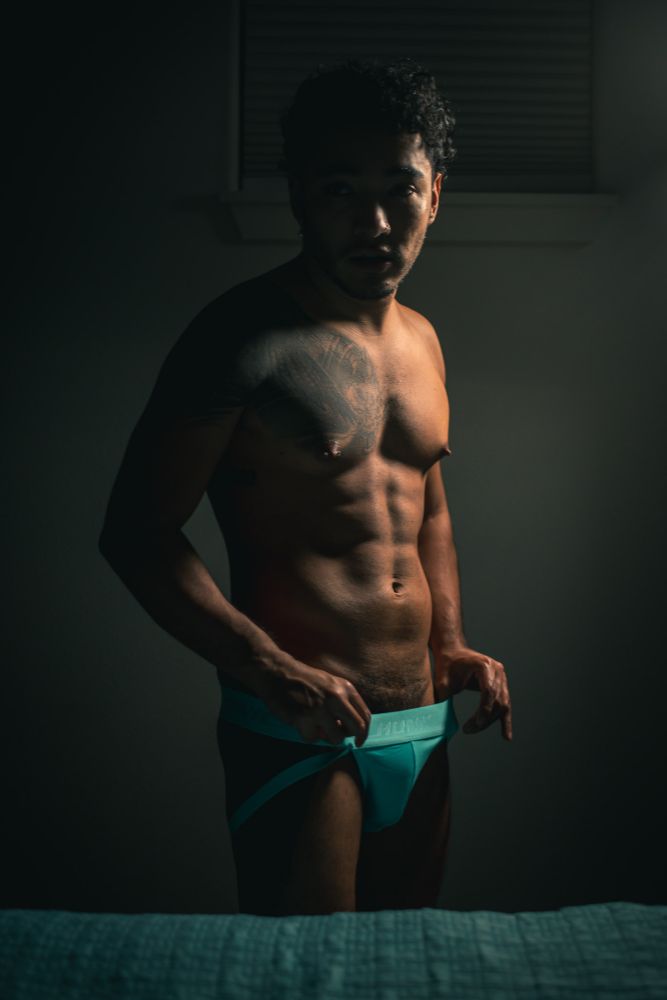 He stands, bedside, in a dimly lit room. Just beginning to tug at the waistband of his turquoise jock strap, he looks to the camera - softly lit from above and right. His abs and chest are on display, a tattoo covering one of his pecs. 