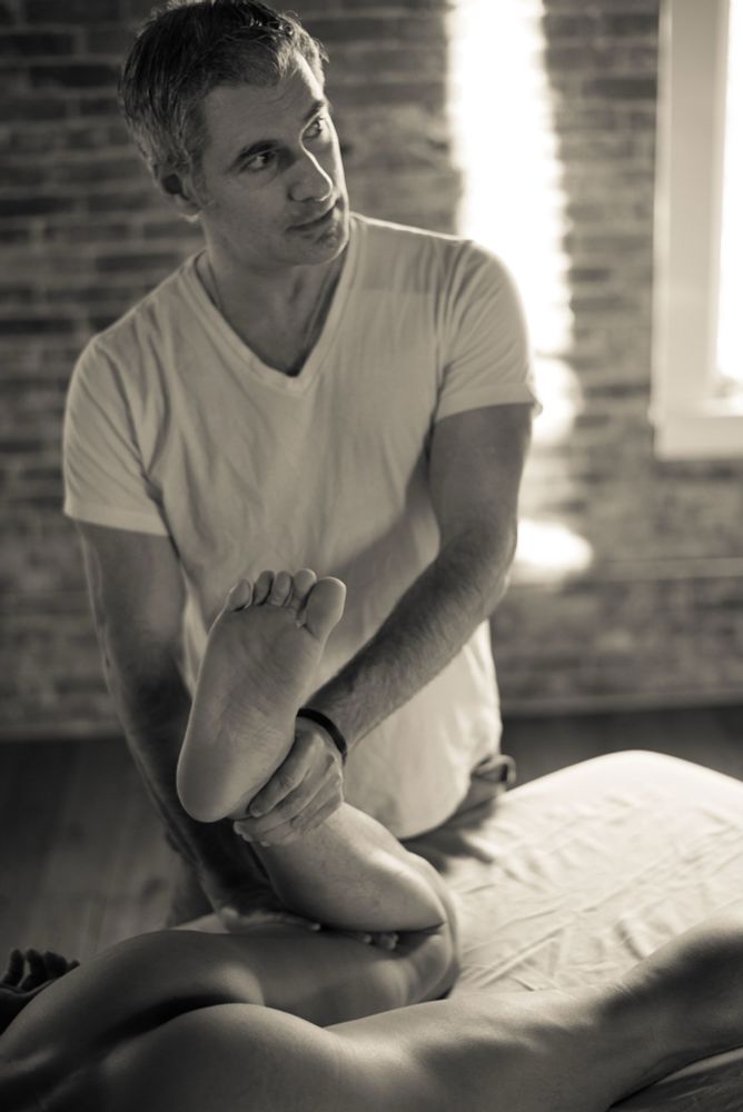 Monochrome image as Joel demonstrates on a nude male model laying face down on a massage table, grabbing his ankle and pressing on the back of his knee - bending the leg. 
