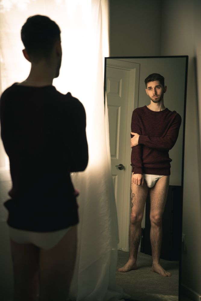 A young man stands in front of a full-length mirror, wearing a maroon sweater and white briefs, with his reflection captured in the mirror. The room is softly lit, creating a moody, introspective atmosphere. He gazes directly into the mirror, holding his left arm with a contemplative expression. A large window with diffused light and a white curtain is visible in the background, adding to the subdued tone of the scene.