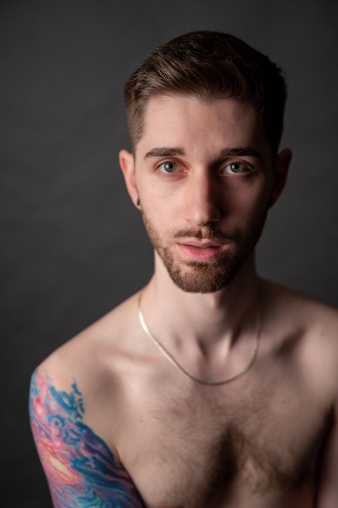 He looks deeply into the camera, a colorful swirling tattoo visible on his shoulder, wearing a simple chain and a dusting of chest hair just in frame