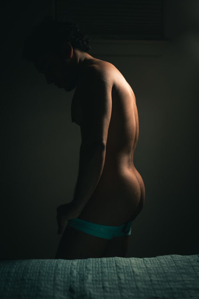 He stands, bedside, in a dimly lit room. Lit from behind, he has pulled down his jockstrap and the light reveals the cheeks of his butt as his face and torso are in shadow